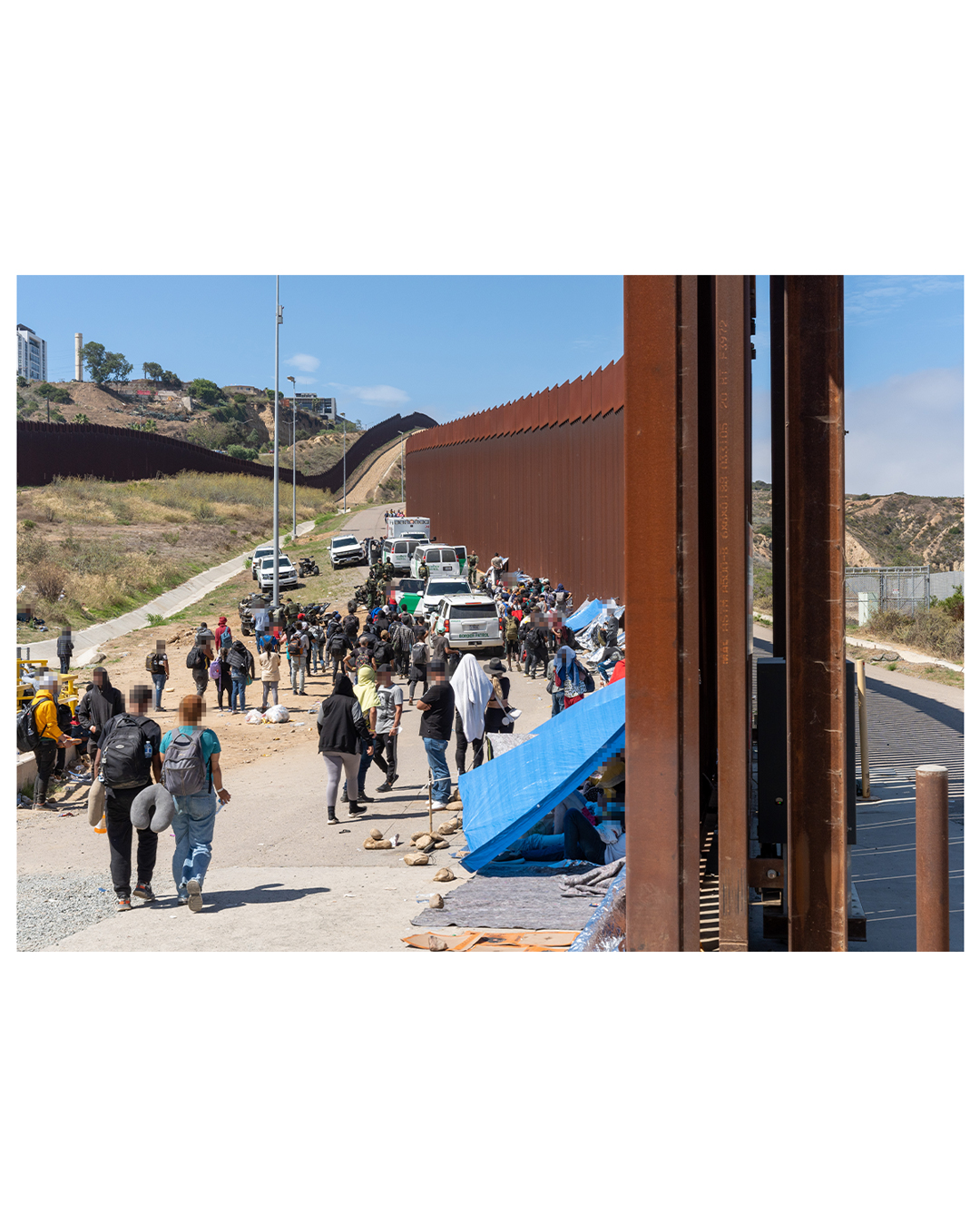 A large group of migrants gathers against the border wall. Some have set up tarps against the slats to shelter from the overhead sun. 7 border patrol vehicles can be seen at the top of the lane, as well as several ATVs. The border patrol agents are noticeably distanced from the migrant population.