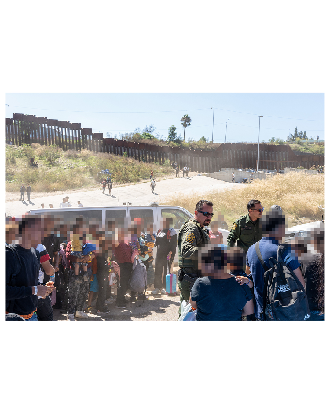 Dozens of migrants stand beside two border patrol vehicles, while two border patrol agents look on. The group of migrants include small children, men, and women. In the background, other migrants can be seen walking near the wall.
