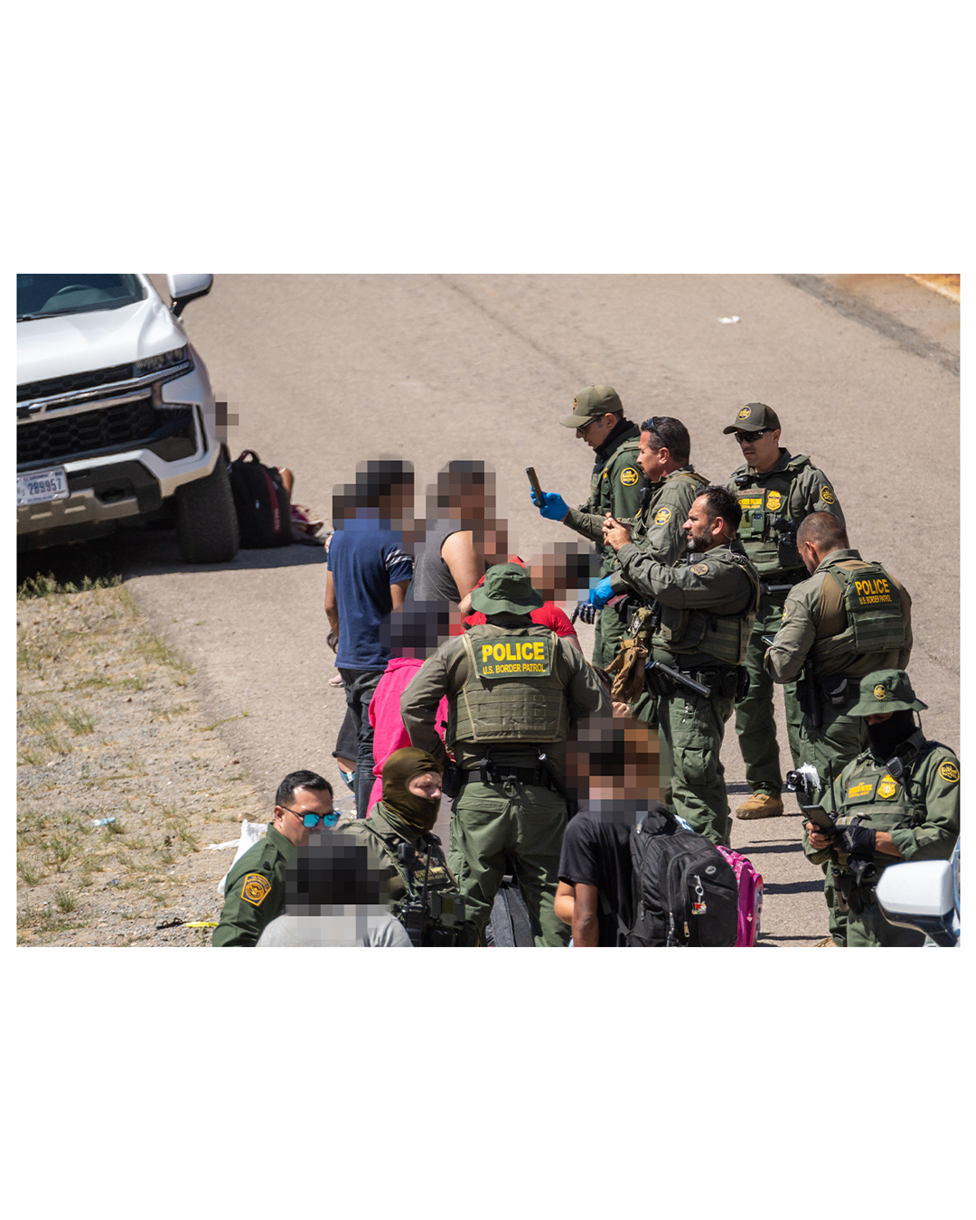 A large group of border patrol agents stand in front of a smaller group of migrants; the agents have phones in their hands as they look over the migrants, some holding the phones as though taking pictures. Several agents wear facemasks.