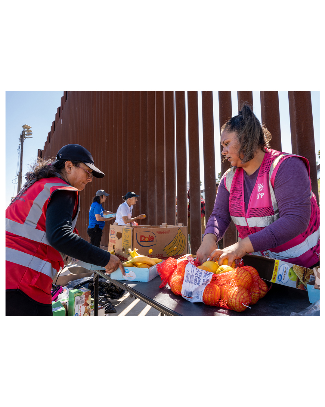 Two volunteers sort through fresh produce on one side of the wall. Behind them, two other volunteers speak to migrants through the slats in the wall, handing out oranges and bananas.
