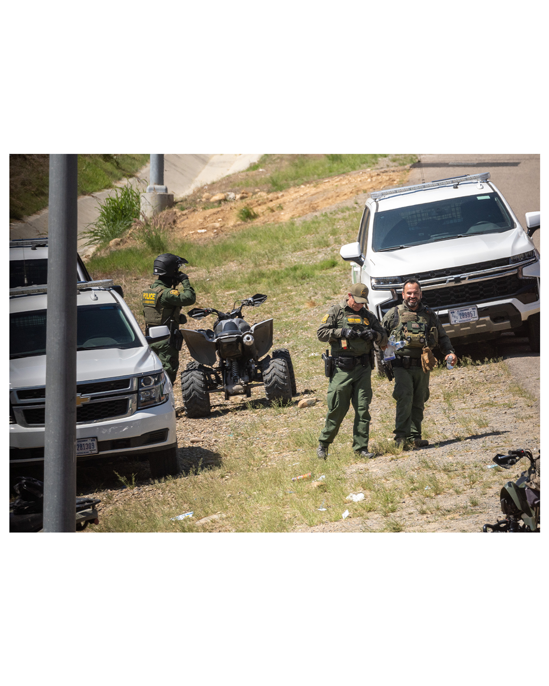 Two border patrol agents walk toward the camera, flanked by border patrol vehicles on both sides. Behind them, a third border patrol agent fiddles with the strap on his helmet as he stands beside a border patrol ATV.