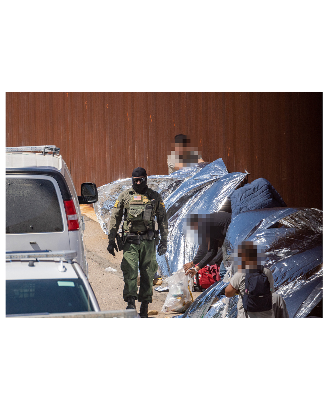 A border patrol agent wearing a ski mask and sunglasses walks along the wall. Tied to the wall are mylar emergency blankets, reflecting the hot midday sun as migrants shelter beneath to escape the heat. A few migrants stand to the side, watching the agent.