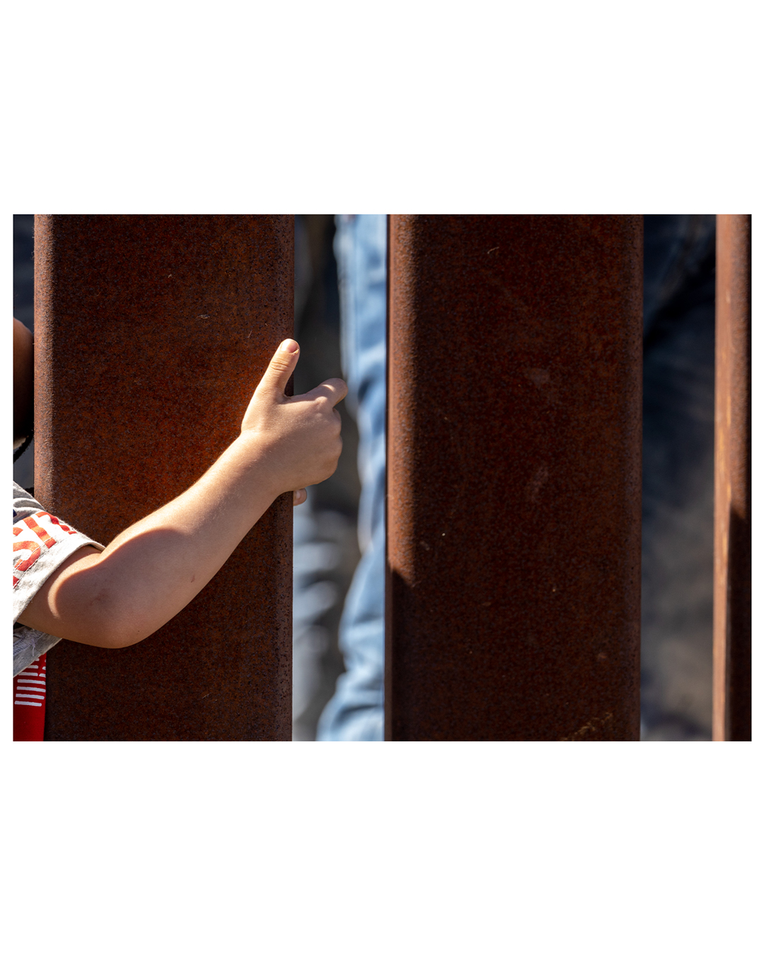 A young person’s arm wraps around one of the slats of the border wall.