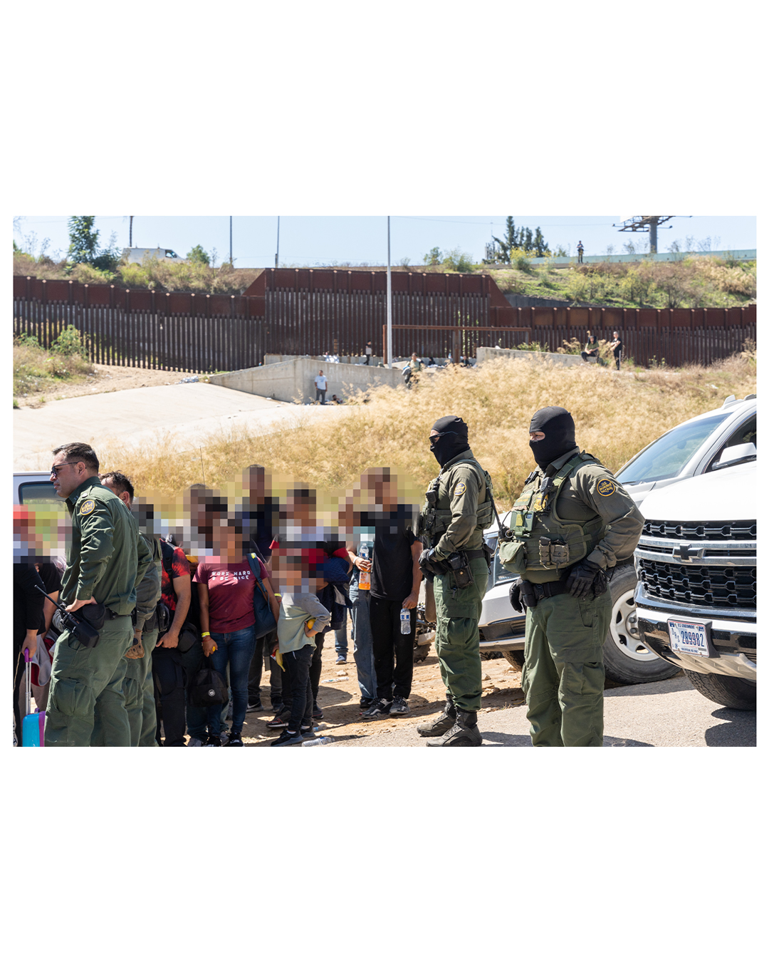 Several border patrol agents stand at the head of a group of migrants. The group includes women and children; they are encircled by border patrol cars while the agents look on.