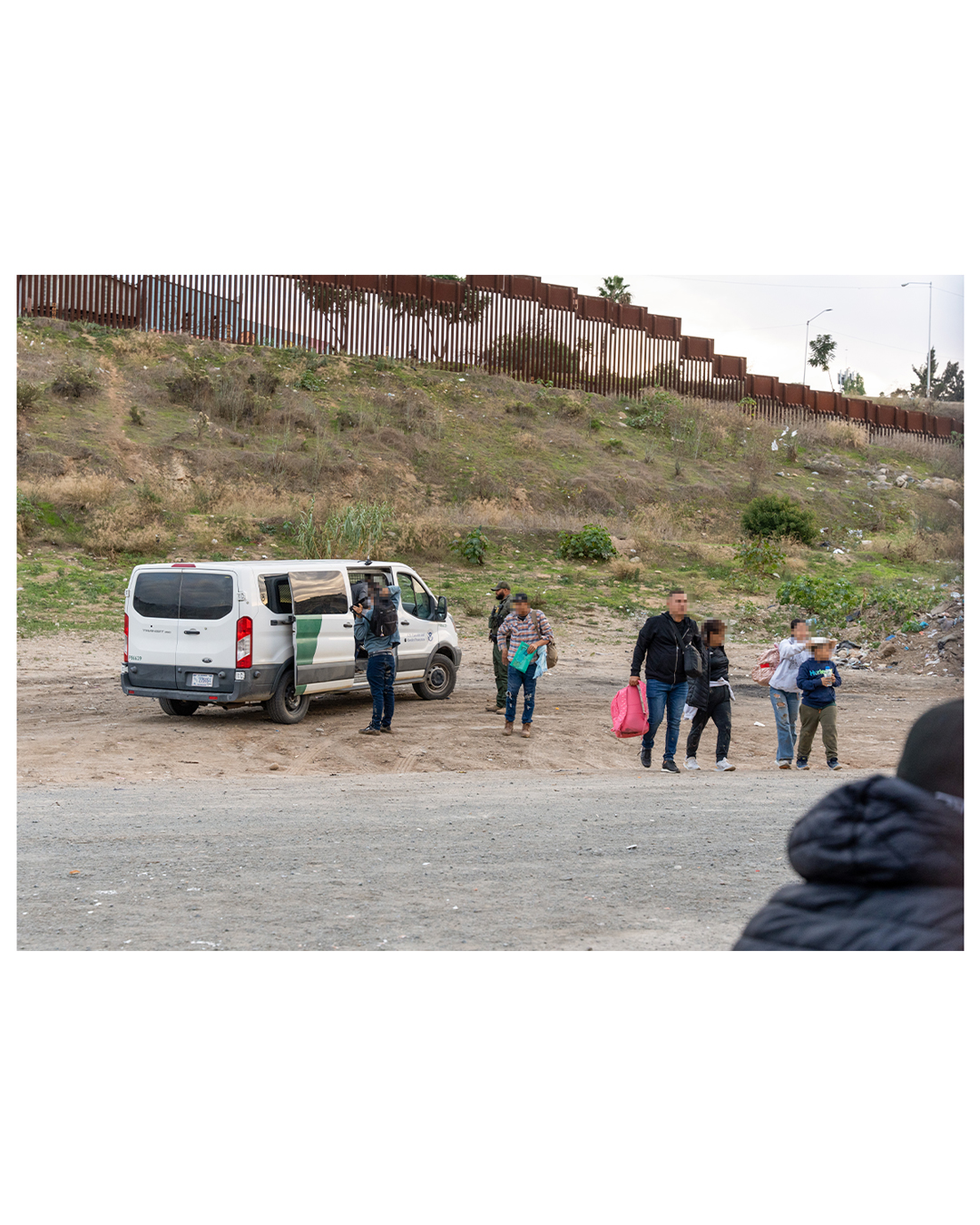 A Border Patrol agent supervises as migrant families leave a vehicle; in the foreground, as four migrants walk away, a woman can be seen adjusting a sunhat on a young child. Each person is carrying nothing but a backpack as they walk away from the van.