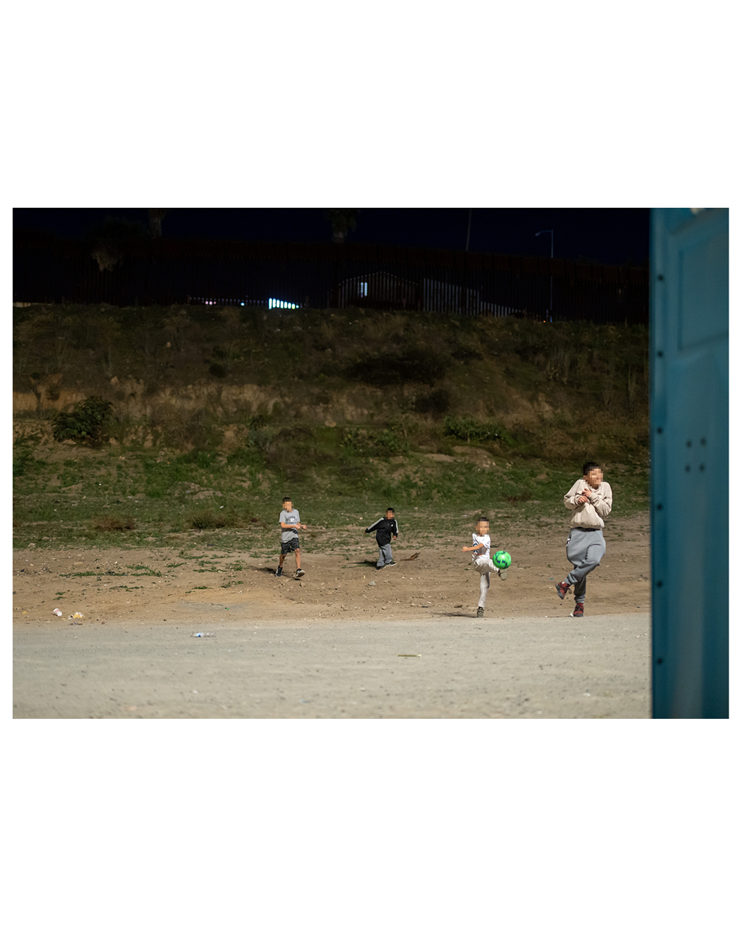 In the dark, four children play soccer together on a long dirt lane. The outline of the border wall is barely visible in the darkness behind them, except a few lights shining through the slats. Trash litters the ground around where they children play.