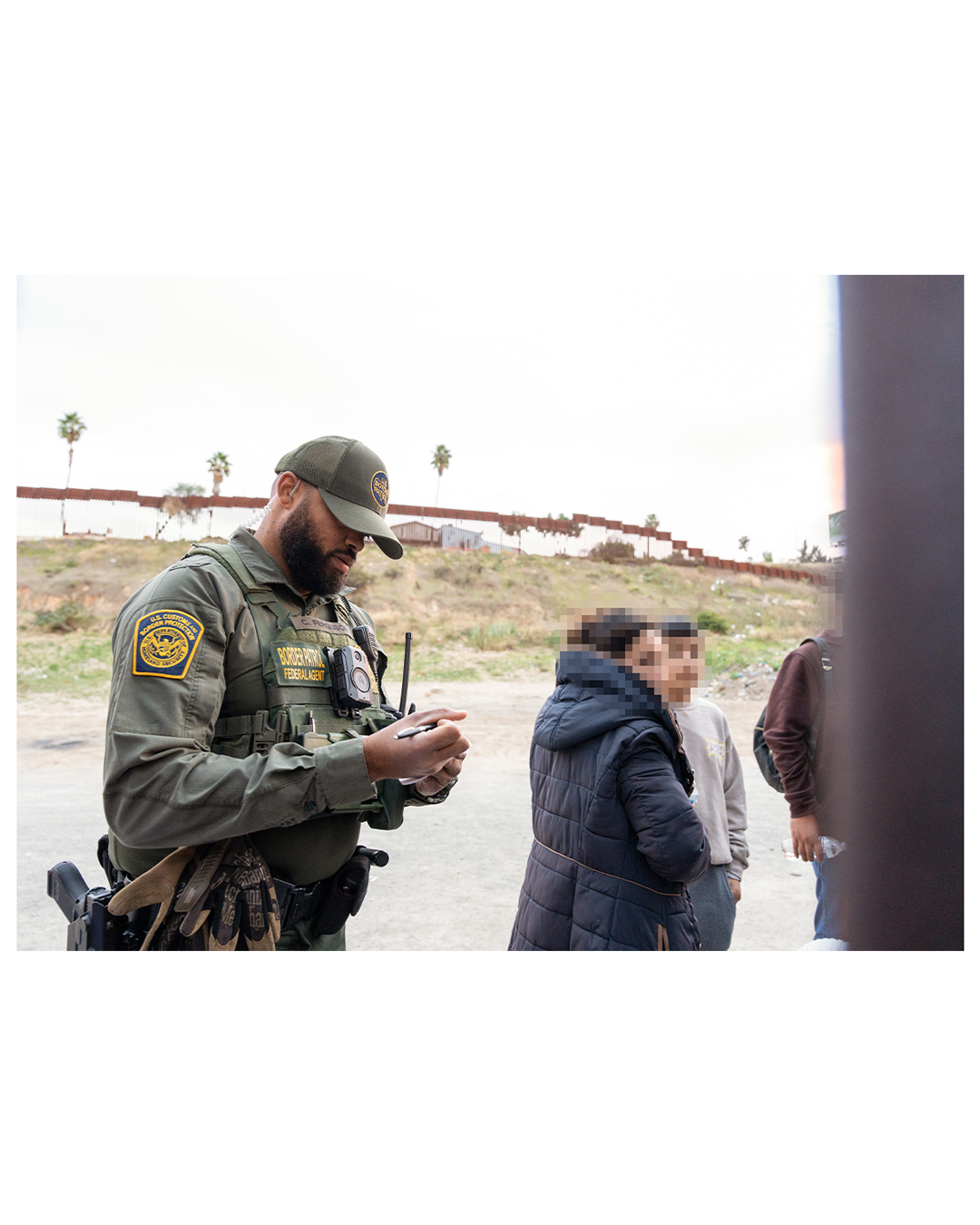 An armed Border Patrol agent holds a pen in his hand as he makes a note. A few migrants stand in front of him. Palm trees and a billboard can be seen in the distant background.