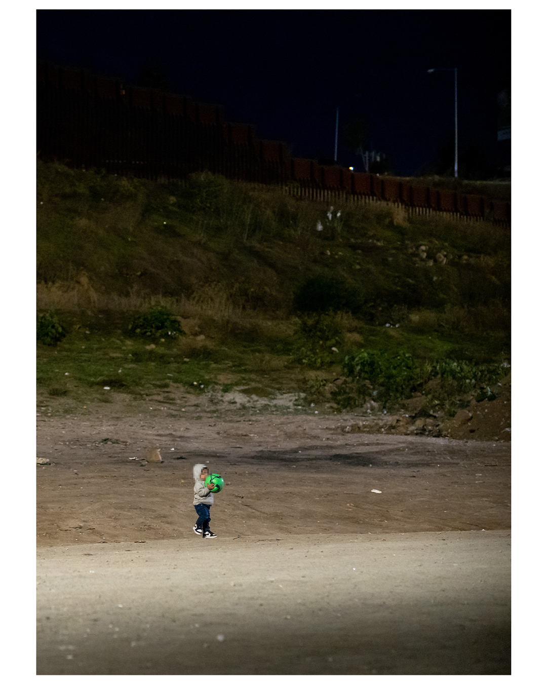 A small boy holds a bright green soccer ball up to his chest, obscuring his face, as he stands in a dirt field at night. Scrub hills and a wall can be made out in the darkness behind him.