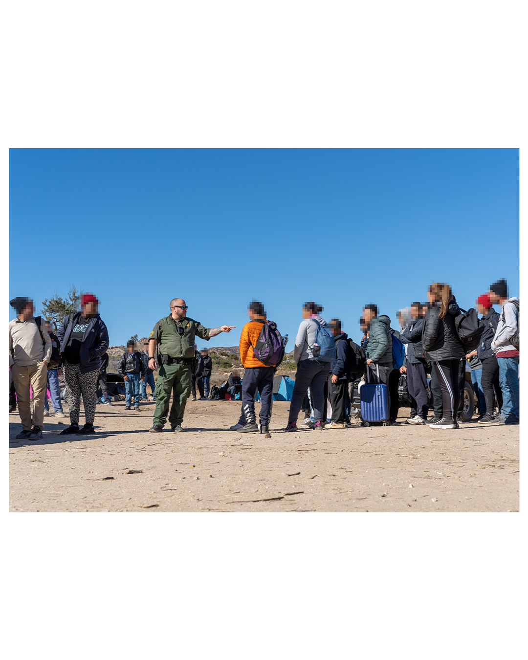 A Border Patrol agent stands in the center of a group; he is pointing his finger directly at one migrant man, while other migrants on all sides of the agent look on.