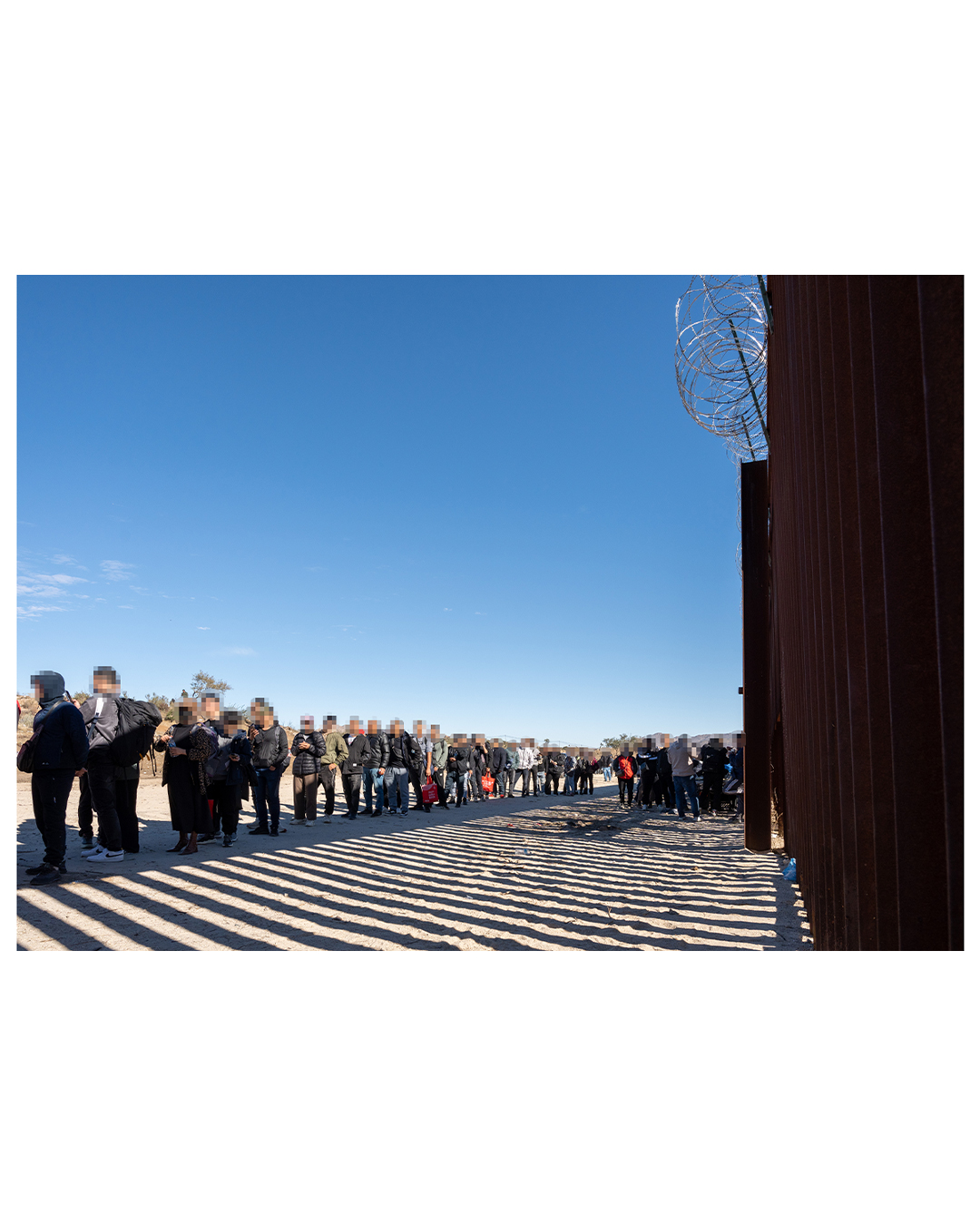 The view from against the border wall. The wall is large slats, through which light shines through, creating a series of light and shadow bars on the ground. A line of migrants stretches as far as the eye can see; a few carry backpacks, most have no belongings in their hands.