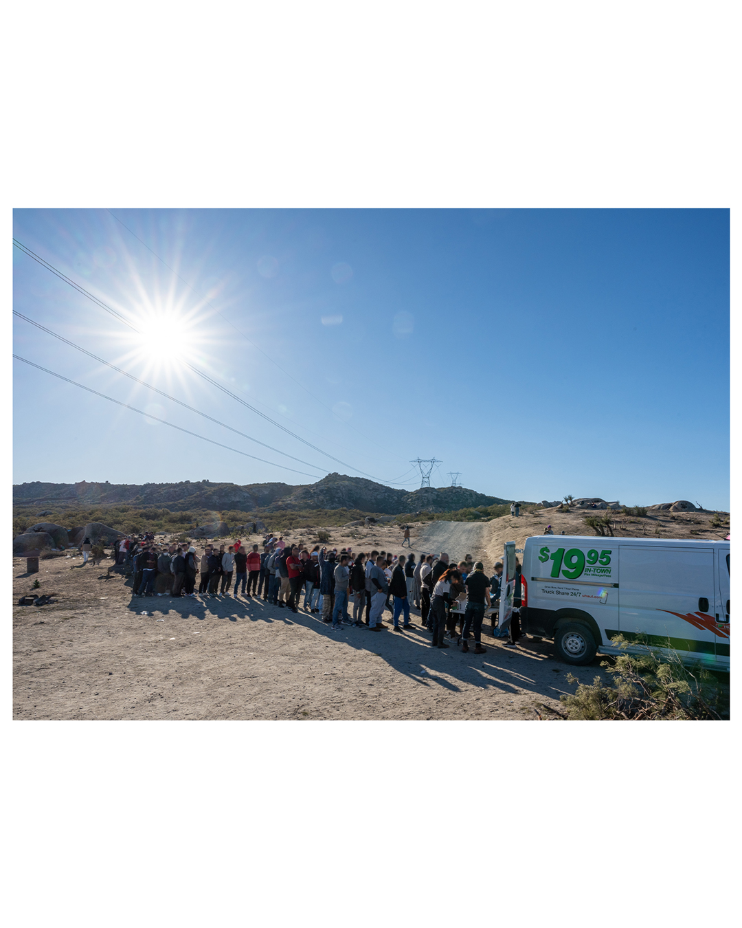 A bright sun shines in the upper left corner. Under the sun, a line of migrants snakes into the distance, as they wait behind a U-Haul truck staffed by volunteers handing out aid. Power lines cross the empty desert and the dirt road the volunteers are parked along.