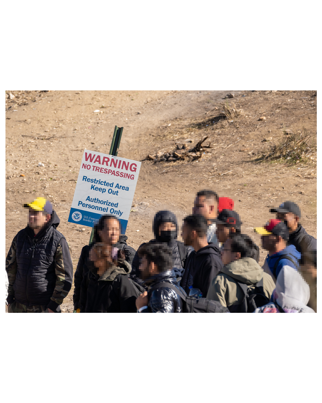 Against an empty desert background is a sign that reads
WARNING: NO TRESSPASSING
Restricted Area
Keep Out
Authorized Personnel Only
In front of the sign is a group of migrant men and women, waiting in the desert sun.