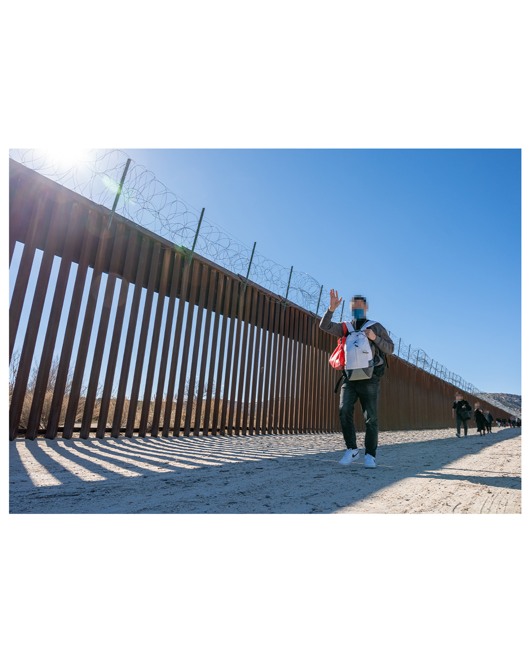 Against the slat metal border wall background, a young migrant waves to the camera. His backpack is worn backwards against his stomach, with a second bag draped over his shoulder. Other migrants walk the same path in the distance.