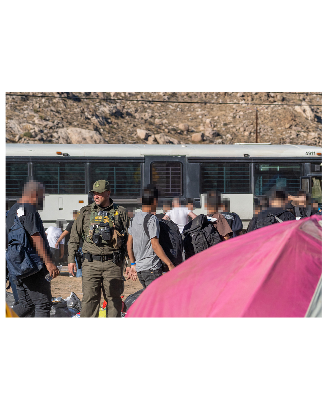 A border patrol agent surveys a group of migrants as they board a bus and load their belongings in the desert; a red tent dominates the lower left foreground. Barren, rocky hills are seen in the background.