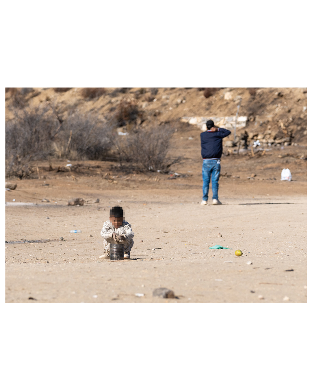 A small boy plays by pouring dirt into a tin can in an open desert field, littered with trash including empty water bottles and a lone lemon. A man in the background has his back turned to the child, his arm up behind his head.