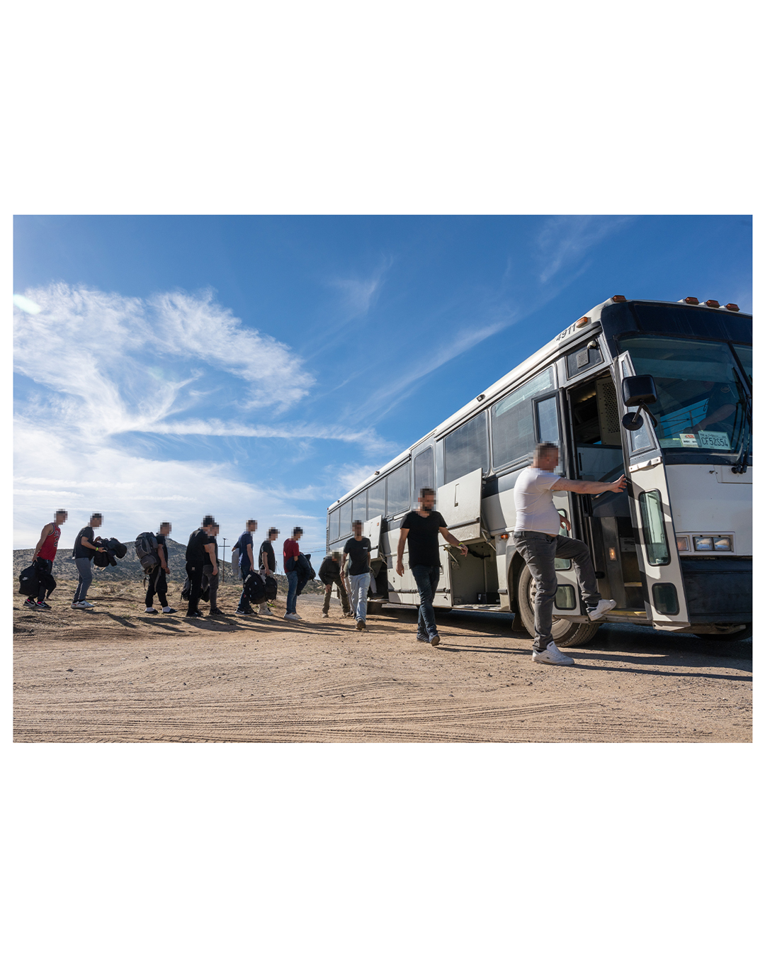 A group of a dozen male migrants line up to board a bus under a blue desert sky after placing their belongings in the bus. Most migrants have only a small backpack containing all their belongings.