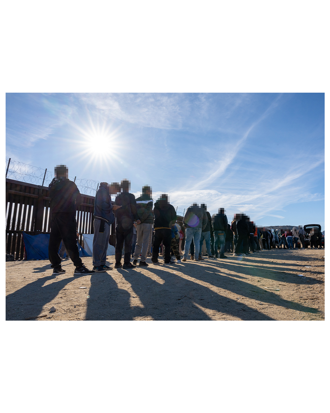 A long line of male presenting migrants lines up against a metal wall topped with barbed wire under a brilliant sun to receive aid from volunteers; the sun dominates the upper right half of the image. Against the wall, a handful of tarps have been set up as makeshift tents. 