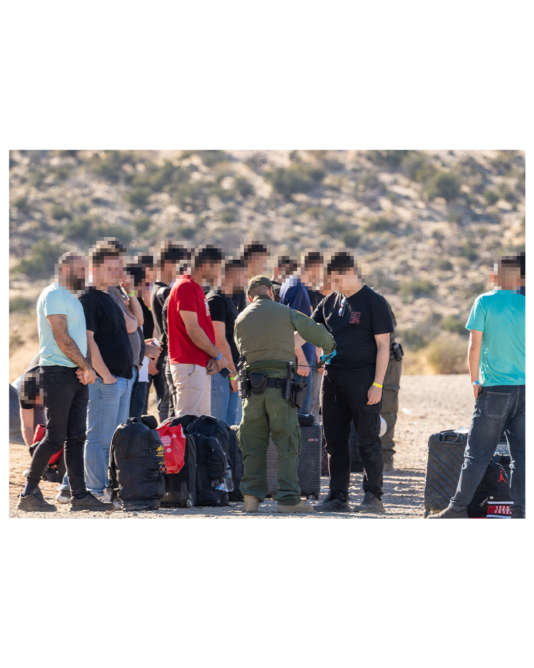 A border patrol agent stands in front of a large group of migrants. In his hand is a set of wire cutters, which he uses to cut the drawstring from the man’s sweatpants. Migrants aren’t allowed drawstrings or shoelaces.. Most of the migrants can be seen wearing either a yellow or a blue wristband. Rocky desert hills dominate the background.