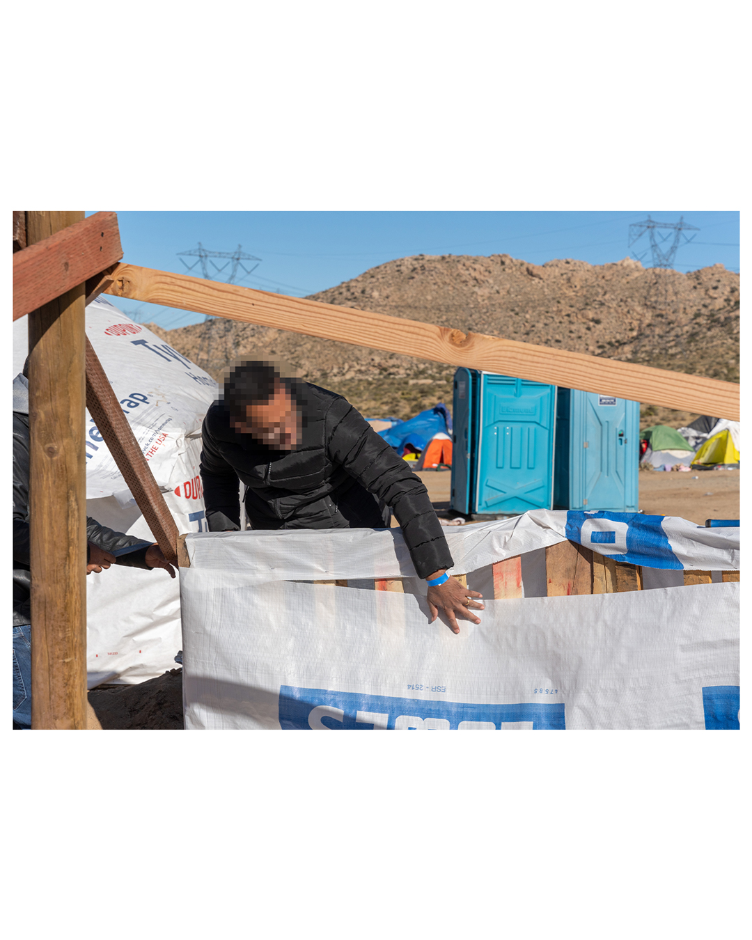 A man leans across the beams of a project in construction to examine a pallet of wood. Behind him are two port-a-potties, and behind that is an encampment of various tents and tarps. The little camp has been set up below a rocky hill; large power lines run across the background.