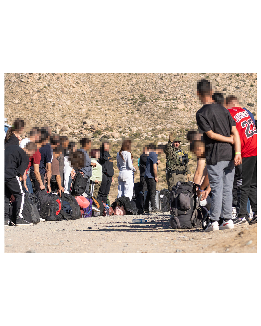 A group of migrants forms a ring around a border patrol agent, his arm raised above his head, palm out and fingers spread. The migrants to the left are all female presenting; to the right, male presenting. Each migrant has a backpack at their feet as they listen to the agent.